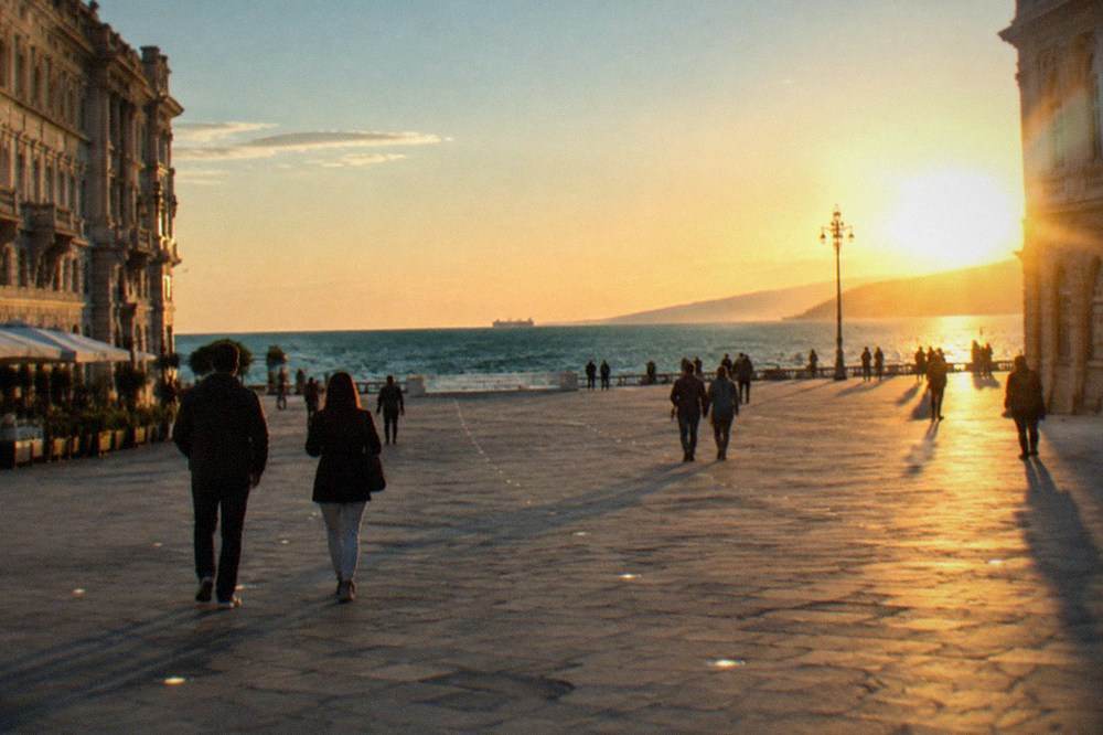 People strolling across a large paved square toward the sea at golden hour
