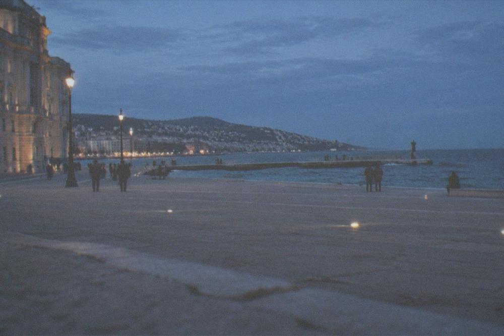 A stone pier extending into calm blue sea with a city skyline behind it