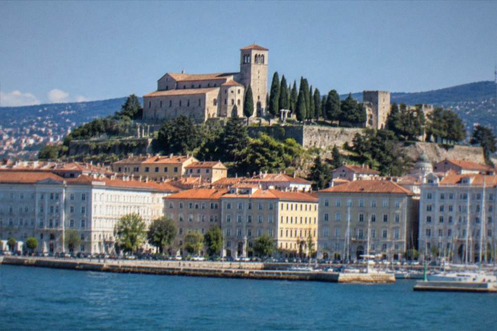 A hilltop castle and cathedral tower seen above the rooftops of an Italian port city