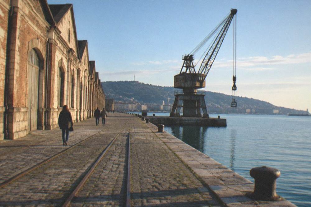 People walking a wide empty quay between old warehouses and the sea