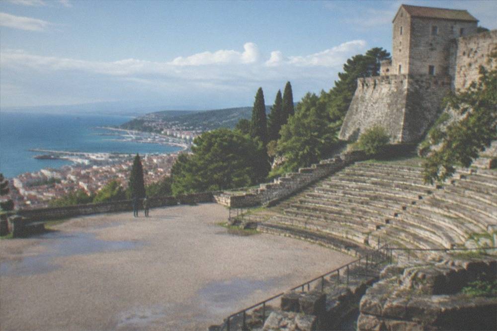 Sunlit stone tiers of the Roman theatre with the green San Giusto hillside rising behind them
