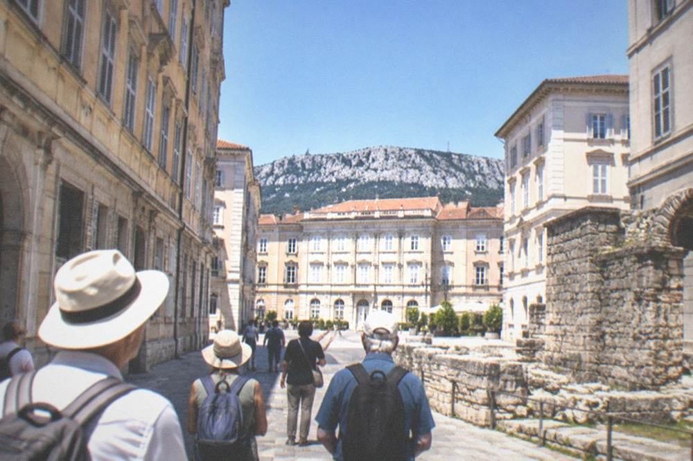 Guided walking group pausing on a narrow old town street near ancient stone ruins in Trieste