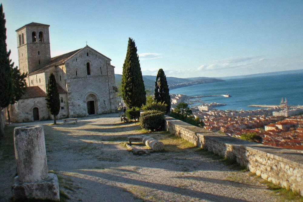 A hilltop cathedral and castle wall overlooking red rooftops and the Adriatic sea