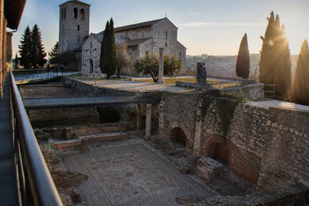 The interior of a brick basilica with a large excavated mosaic floor visible below a walkway