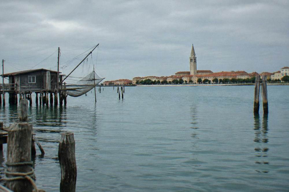 A bell tower rising over red rooftops seen across a calm lagoon