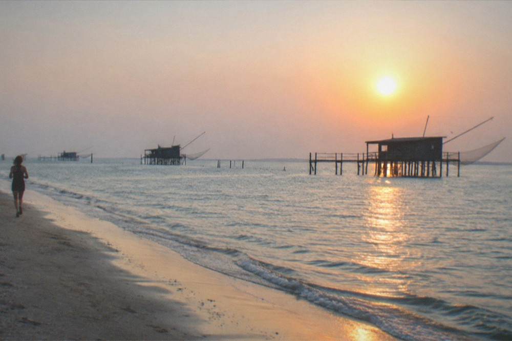 A sunset over a calm lagoon with silhouetted wooden fishing huts on stilts