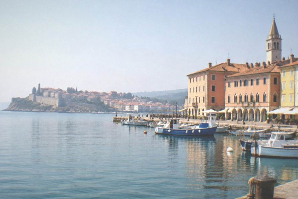 A walled seaside town on a promontory seen across calm Adriatic water