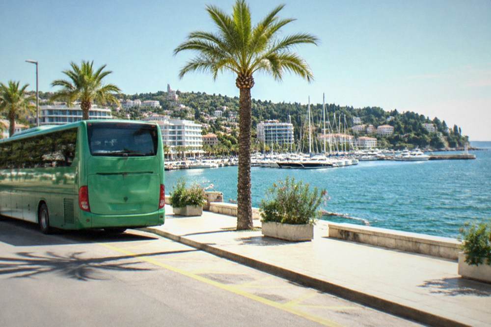 A green regional coach parked at a coastal station with the sea behind it