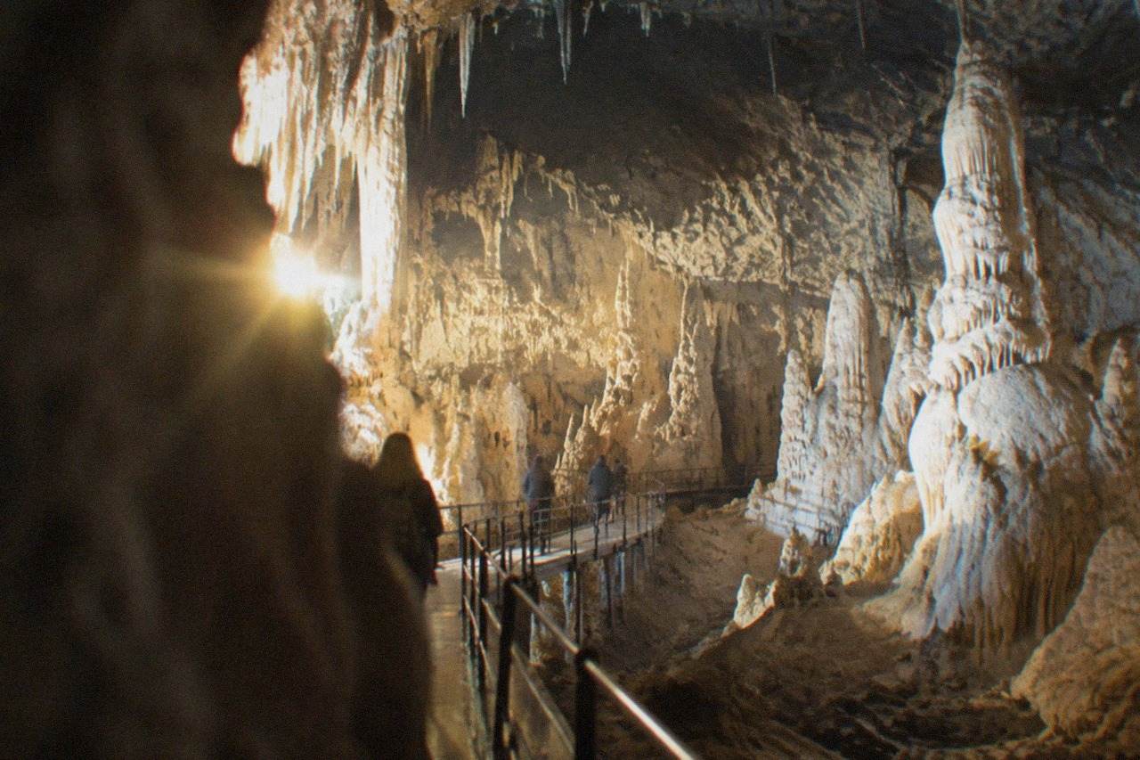 A narrow footbridge curving through a vast illuminated limestone cave chamber with stalactites overhead