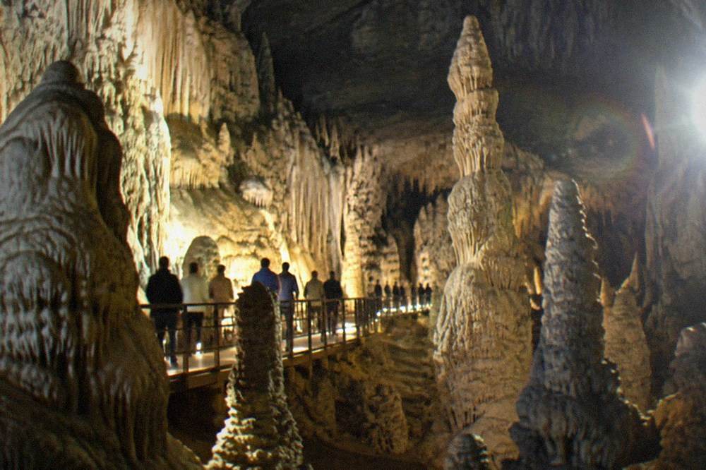 A tour group walking along a lit path between tall stalagmite formations inside a limestone cavern
