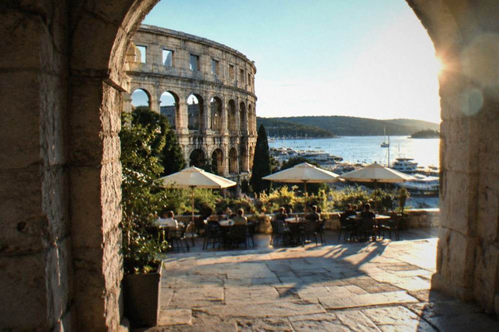 A stone archway in an old Croatian town with cafe tables in the square beyond it