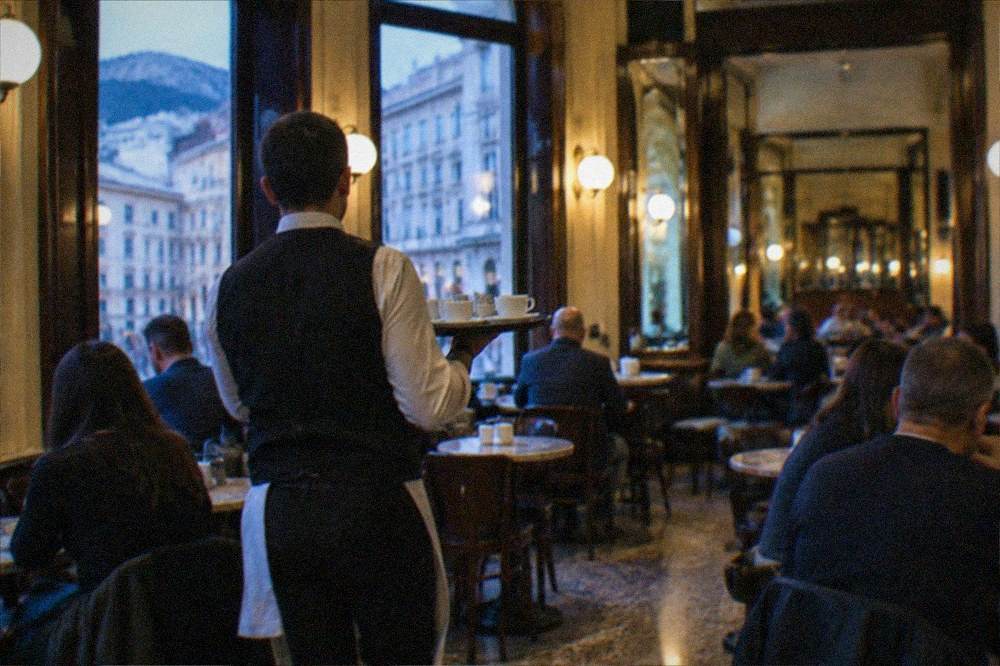 A waiter carrying a tray of coffee cups through a busy old cafe dining room