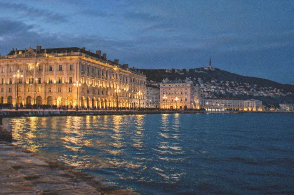 An evening view of the Trieste waterfront with the lit facade of a grand square facing the dark sea
