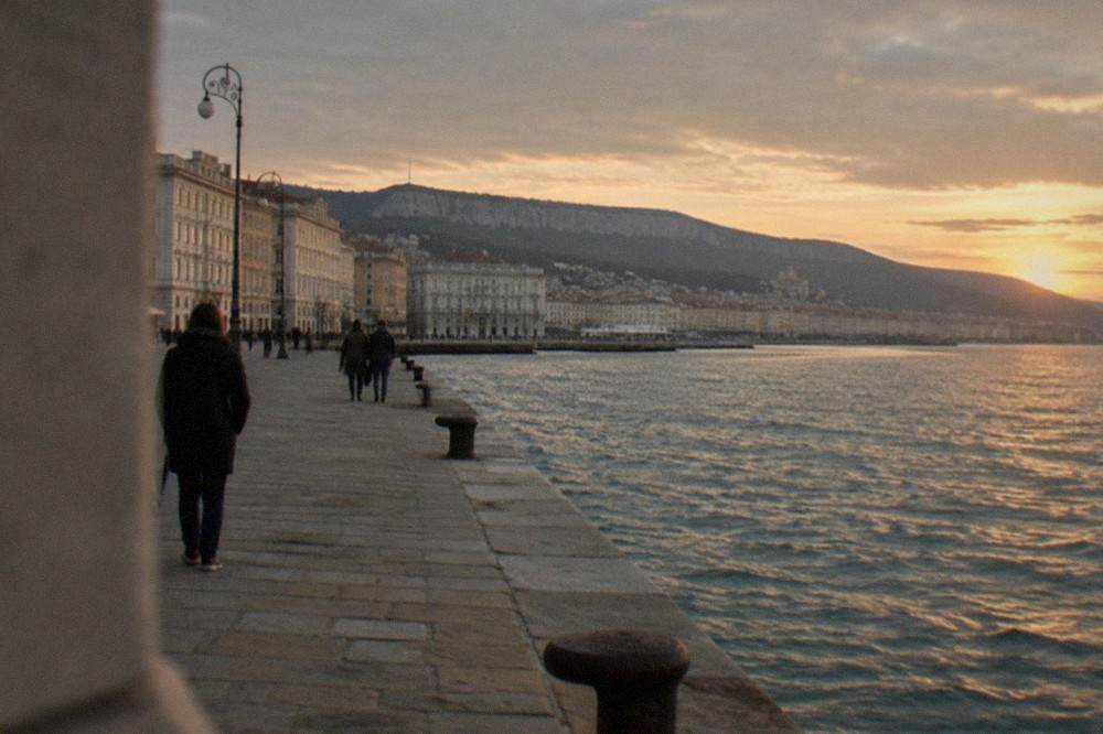 People strolling on a stone jetty reaching out over calm Adriatic water at golden hour