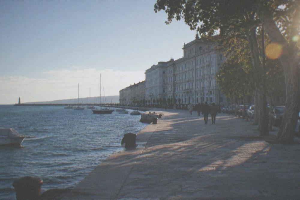 A long stone seafront promenade curving along a blue gulf with a distant lighthouse on the point