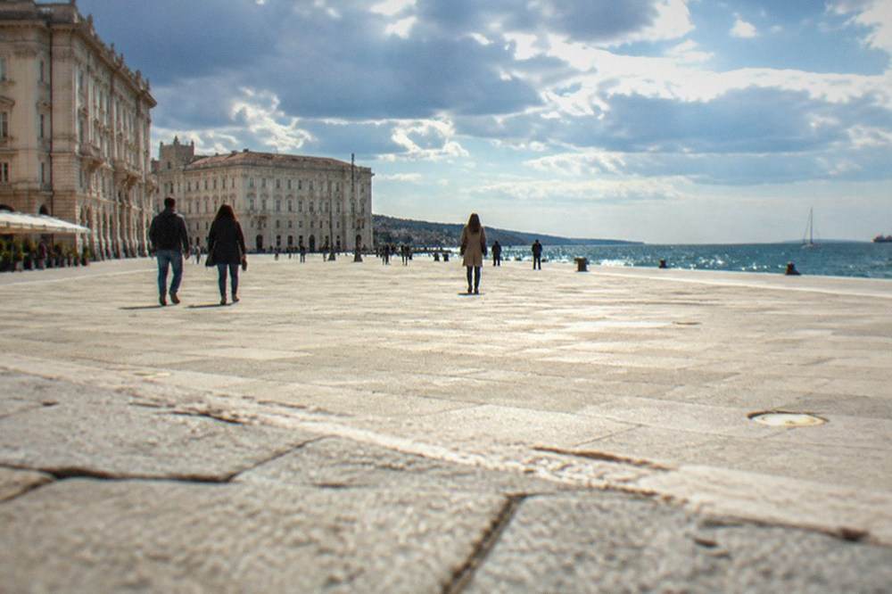 Walkers crossing the vast open seafront square Piazza Unità d'Italia in Trieste with the sea on one side