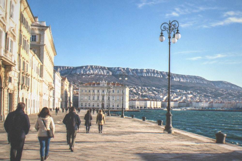 A walking group in light jackets passing grand seaside buildings under a windy clear sky in Trieste