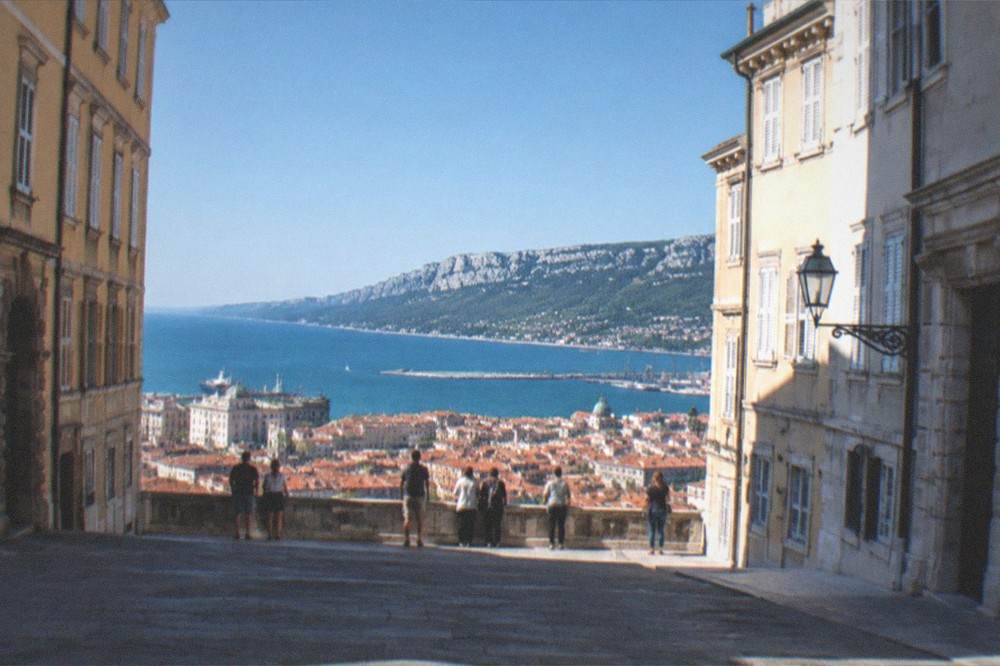 A walking group pausing at a viewpoint over the red rooftops of Trieste and the Adriatic sea beyond