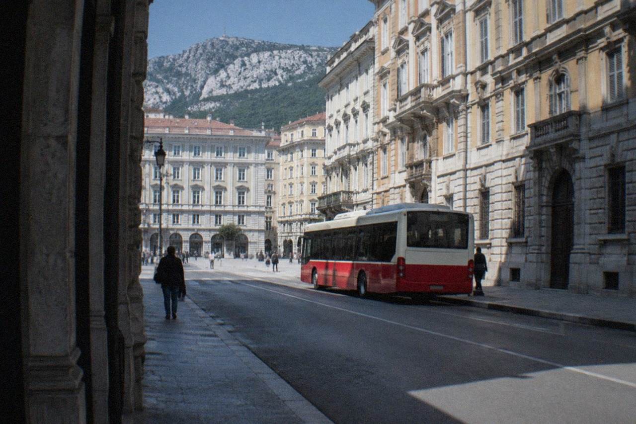 A red and cream city bus stopped on a Trieste street with Habsburg-era buildings behind it
