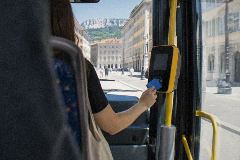 A passenger tapping a contactless bank card on a ticket reader inside a Trieste city bus