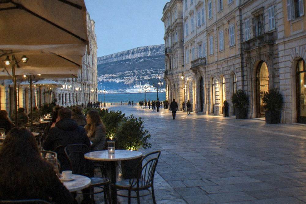 Lit terrace of a historic Trieste caffè at night with people seated at small marble tables