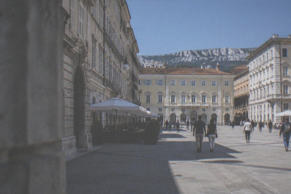 Wide view of a bright Trieste square with locals walking, outdoor cafès and historic facades