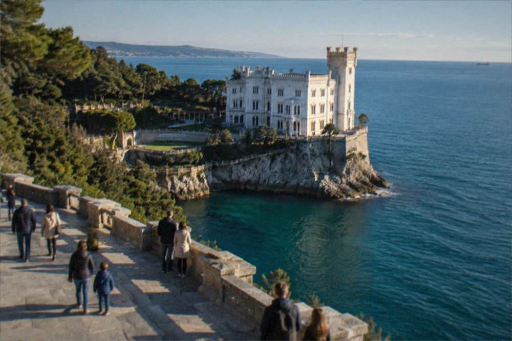 Families walking on a wide stone terrace overlooking Miramare Castle and the Adriatic