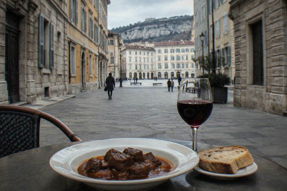 A plate of dark beef goulash with a slice of bread beside a glass of red Terrano wine