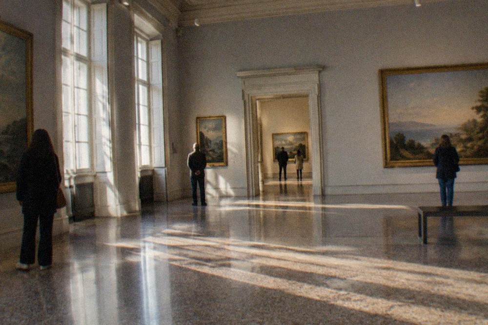 Gallery visitors looking at large paintings in a high-ceilinged 19th-century museum hall