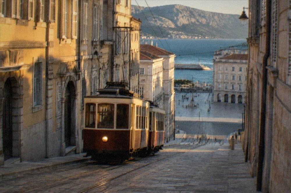A historic two-car tram climbing a hill street above a coastal city