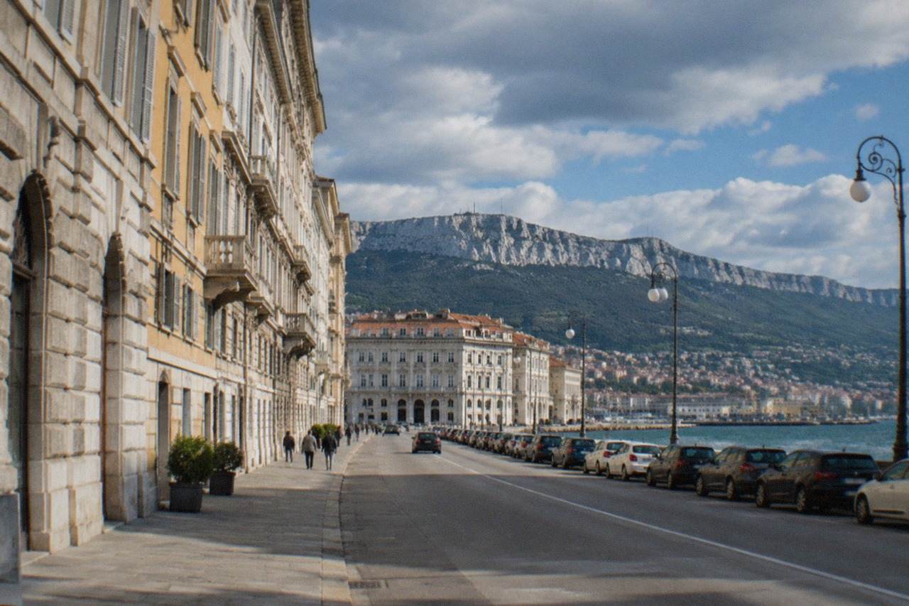 Cars parked along a wide seaside street in Trieste with neoclassical buildings and the Adriatic sea in the background