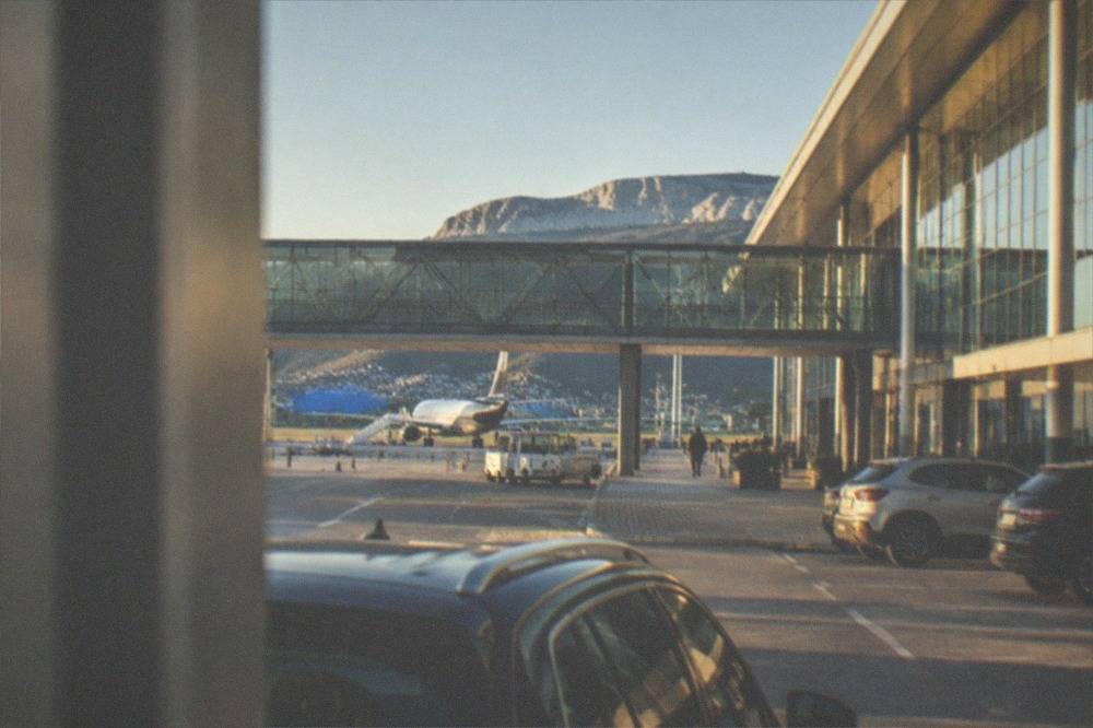 A modern airport terminal building with a covered walkway bridge and parked cars in the foreground