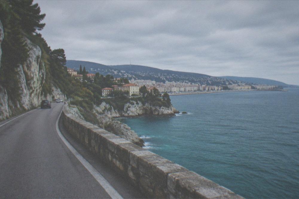 A coastal road winding along cliffs above a turquoise sea with a small town in the distance