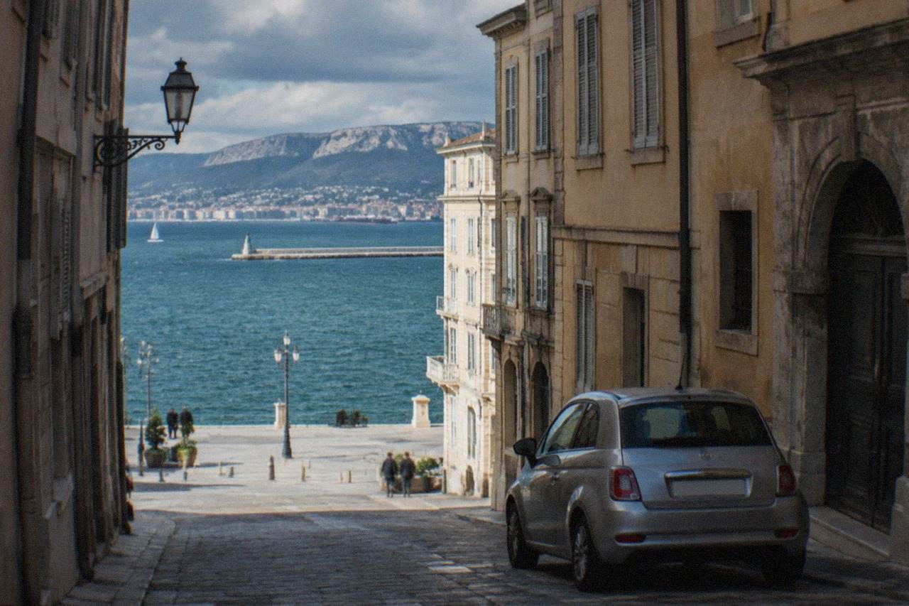 A small rental car parked on a narrow street above the seafront of Trieste with the Gulf of Trieste in the background