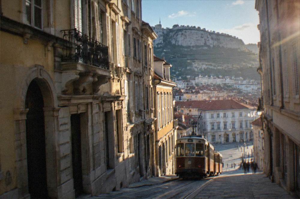 A vintage two-carriage tram climbing a hill above red rooftops toward a limestone plateau