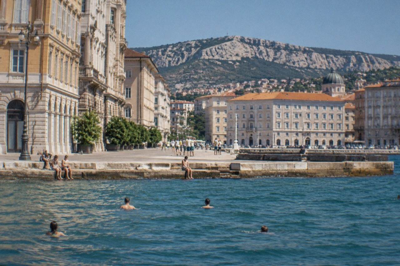 Swimmers in the sea along the Trieste seafront with the city's waterfront buildings behind them