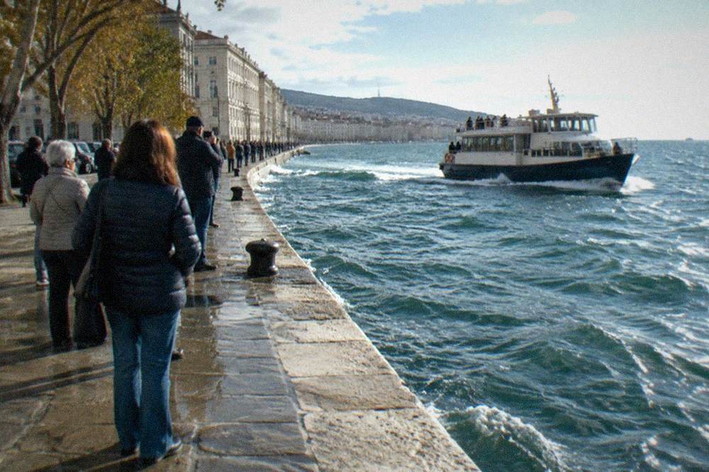 Crowds in coats and sunglasses standing along a stone seafront promenade watching boats pass close to shore
