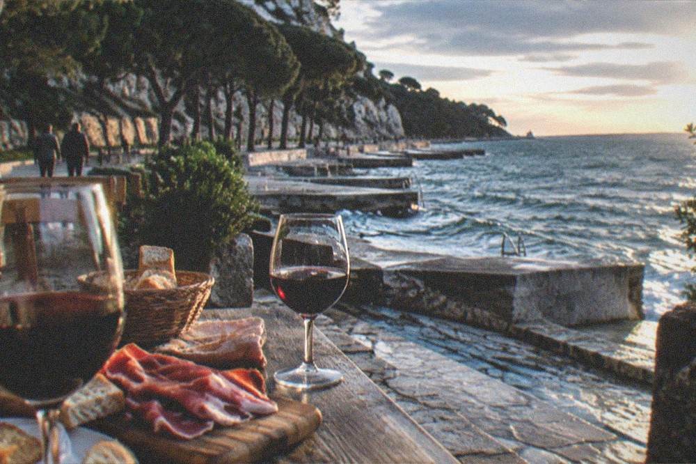 A wooden farm tavern table set with glasses of red wine, bread and cured ham, in a rural hillside courtyard