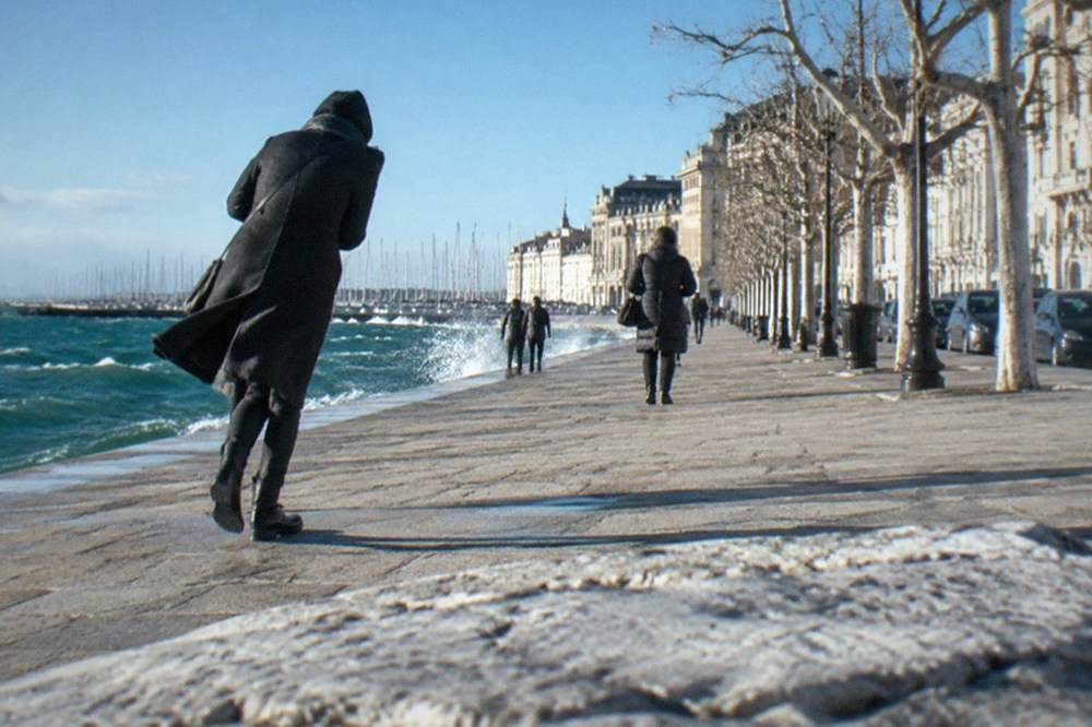 A person leaning into the wind on a wide seafront promenade, coat pressed flat by the gusts