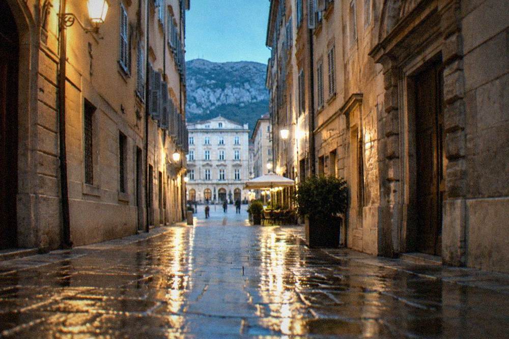 A narrow lane in the Trieste old town at night with lit bar signs and people standing with drinks