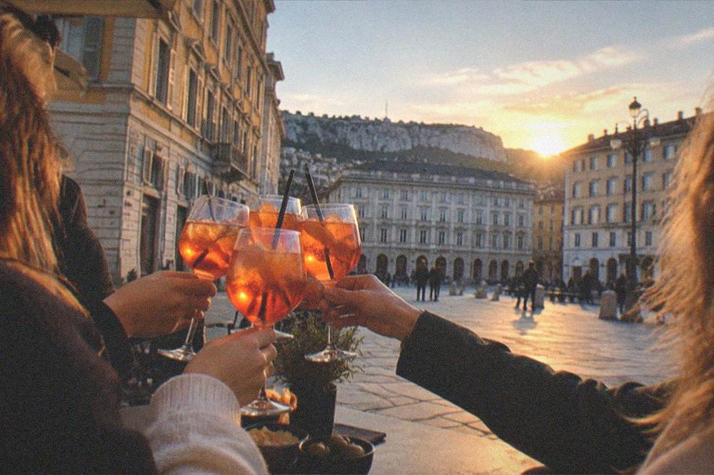 A group of friends toasting with spritz glasses at an outdoor table in a Trieste square at dusk