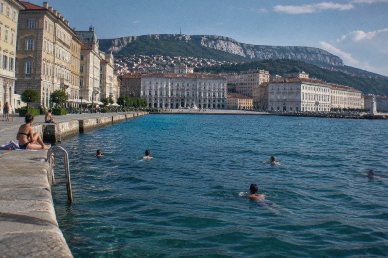 Swimmers in the Adriatic sea beside a stone jetty on the Trieste waterfront with the city behind