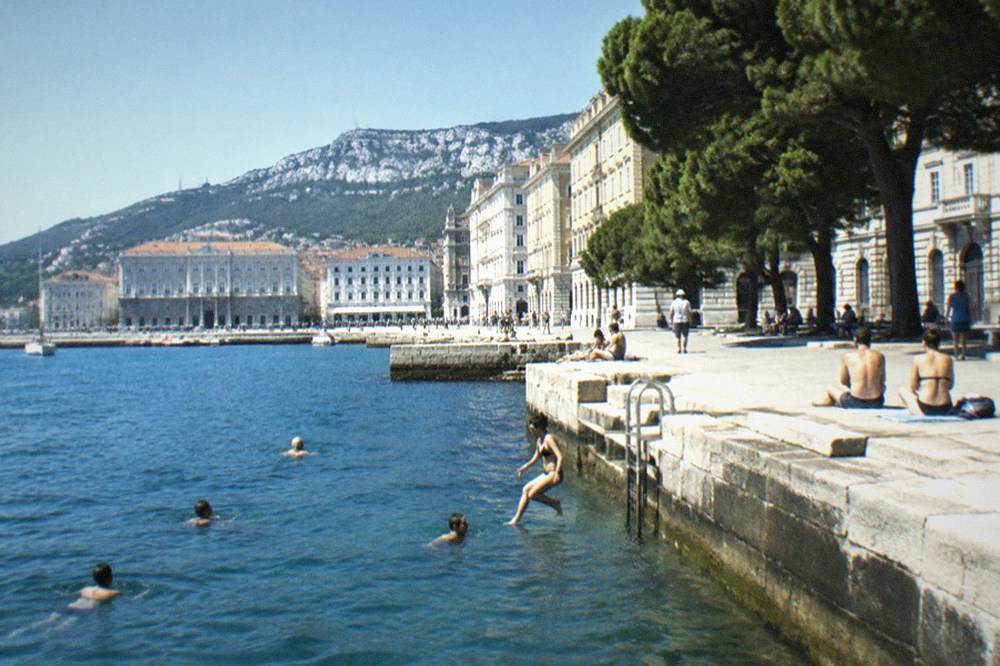 People swimming from a stone platform into calm blue water at the Trieste seafront in summer