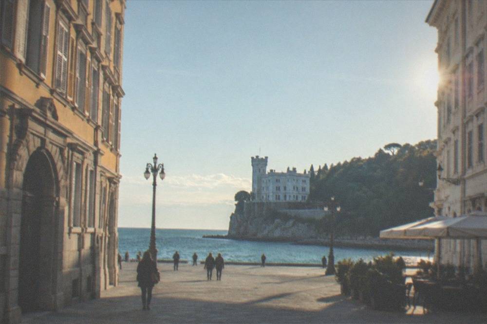 A white castle on a rocky headland above the sea with a wooded park behind it