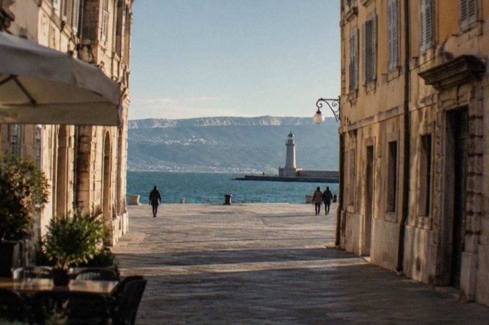 A stone jetty pointing into a calm Adriatic gulf with a lighthouse and distant hills on the horizon