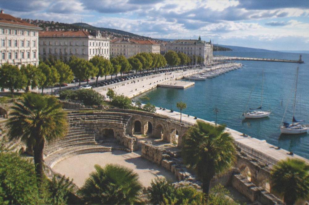 A Roman amphitheatre beside a blue harbour with palm trees in the foreground