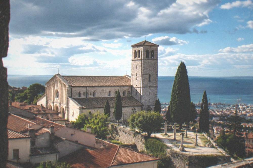 A cathedral facade and bell tower above old rooftops with the sea on the horizon