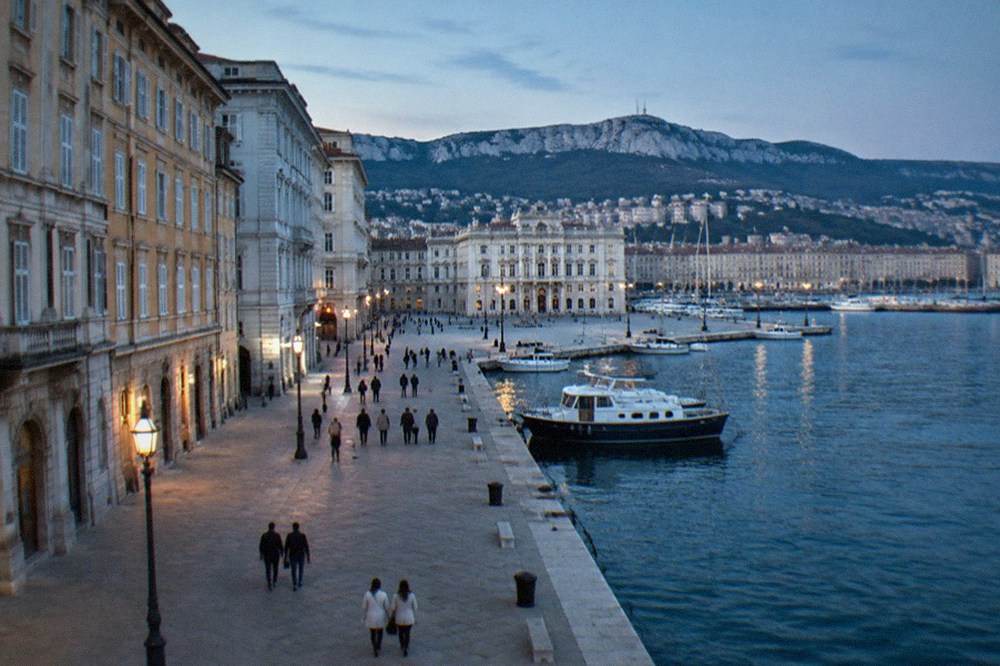Passengers strolling along the Trieste seafront with the harbour on one side