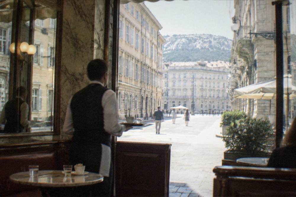A marble café interior with mirrored walls, wooden booths and a waiter carrying a small coffee tray