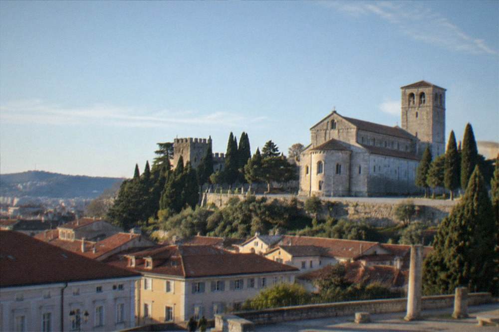 A hilltop castle and cathedral tower above red rooftops seen from a city square